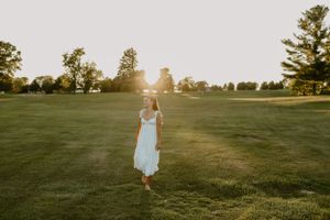 Figure in a white dress walks through a sunlit grassy field at golden hour.