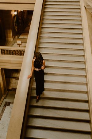 Person in black outfit ascending large marble staircase in vintage-style building.