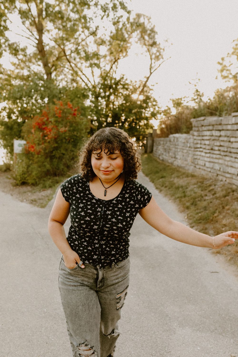 Person in polka dot top and jeans dancing on a path near stone wall.