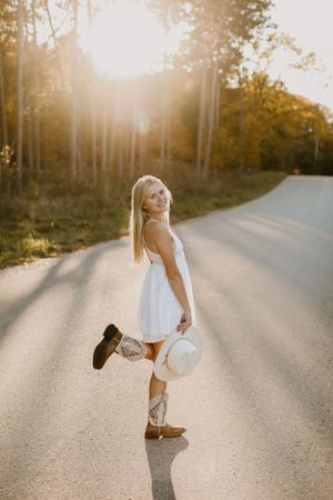 Someone in a white dress poses playfully with boots and a hat on a sunlit country road.
