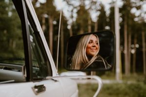 Person smiling from inside a vehicle window surrounded by trees.