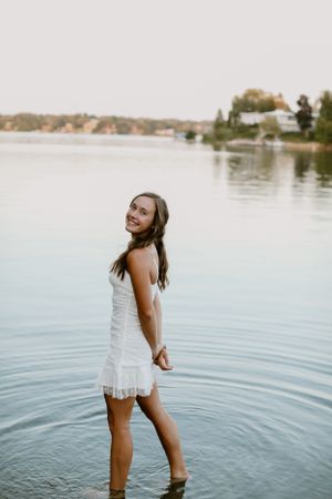 A woman in a white dress stands in shallow water at the edge of a serene lake.