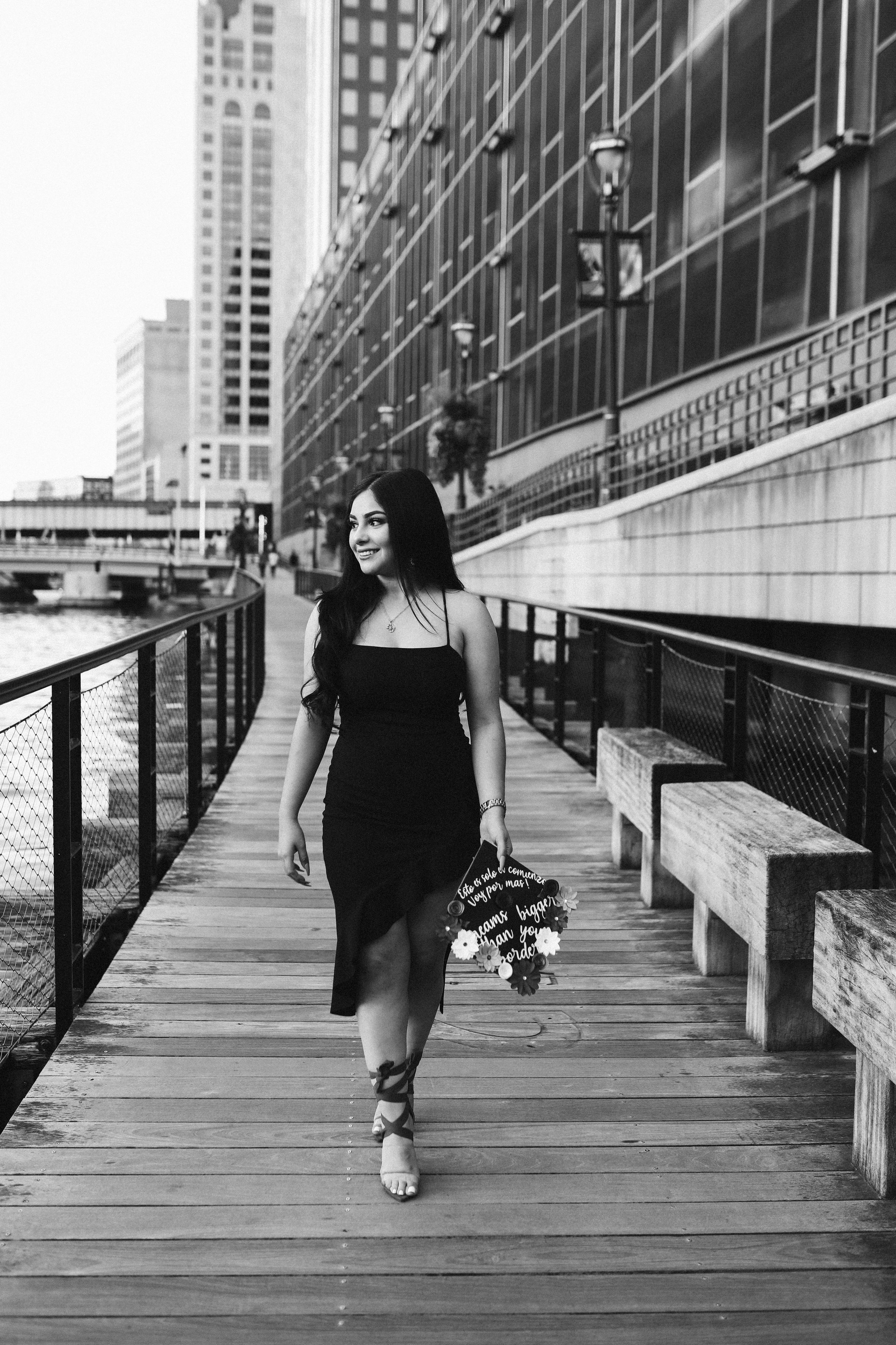 Woman in black dress walks along urban waterfront boardwalk carrying roller skates.