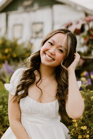 Person in white dress posing in a garden with colorful flowers and house in background.