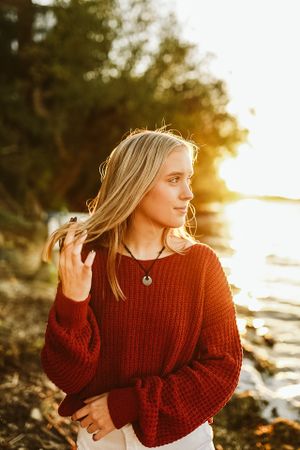 Young woman in red sweater poses at sunset by the water with golden light illuminating her hair.