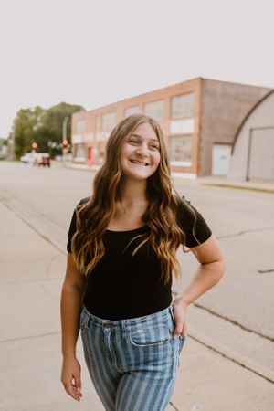 Young person wearing black top and striped jeans standing on sidewalk near brick building.