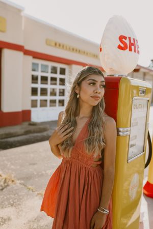 Person in orange dress poses next to vintage gas pump outside retro building.