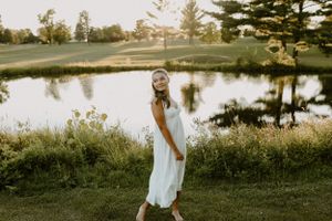 Person in flowing white dress twirls playfully by pond with trees in background.