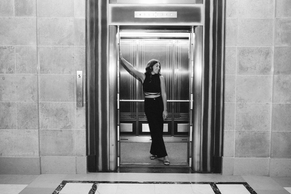 Person posing dramatically in black outfit inside elevator in black and white photo.