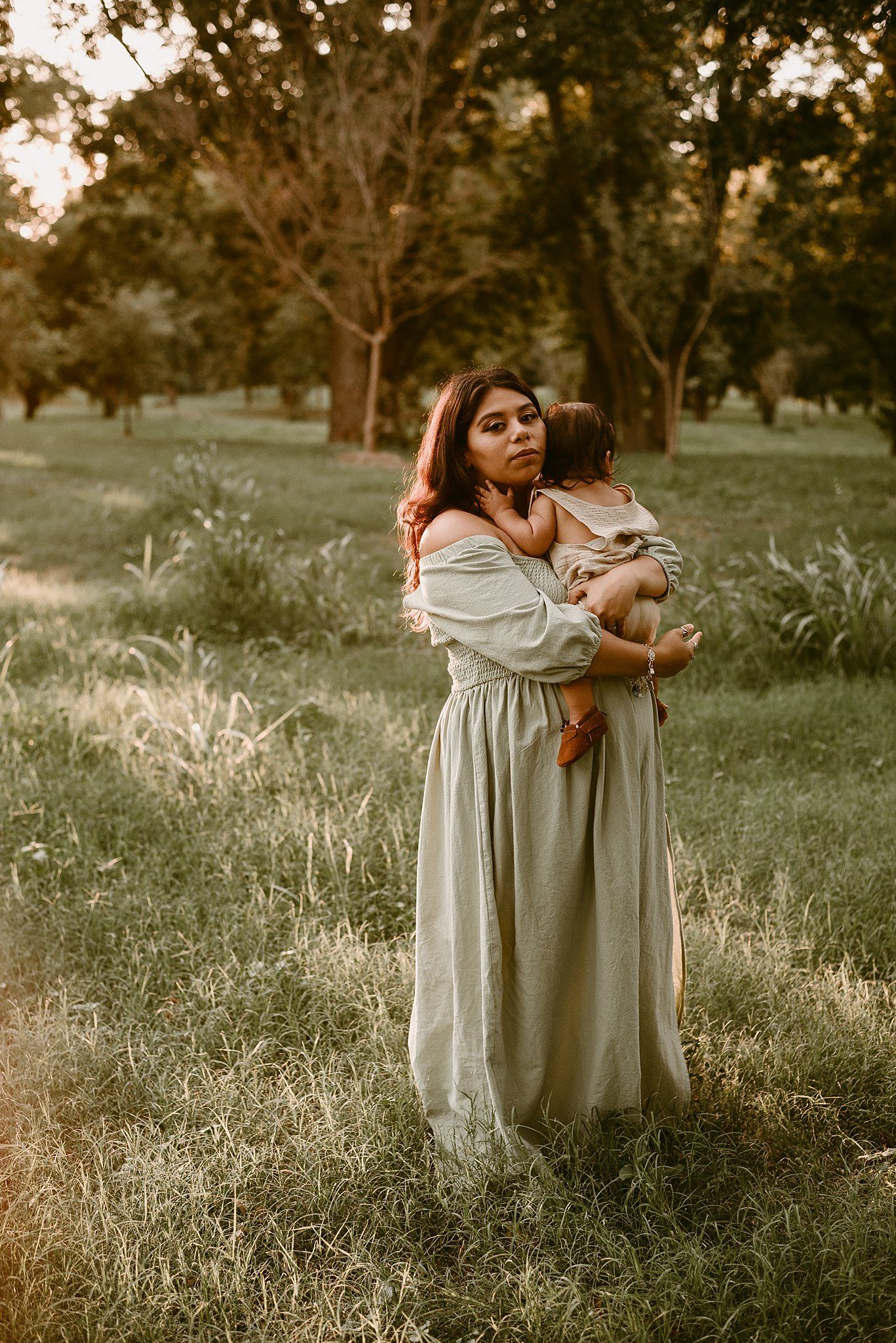A tender outdoor portrait of a woman in an off-shoulder dress embracing a small child at sunset.