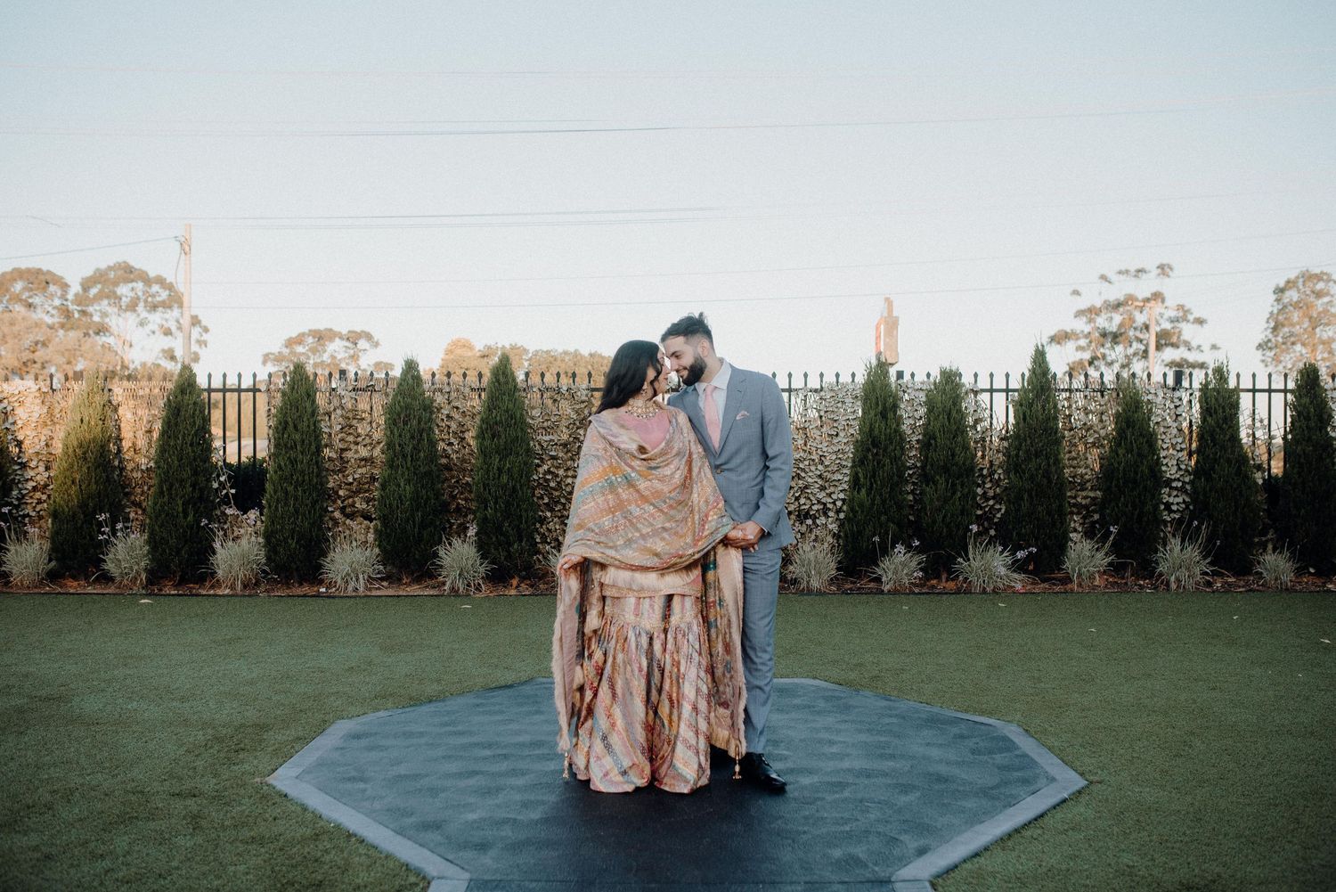 Couple in elegant wedding attire standing on blue platform in garden setting.