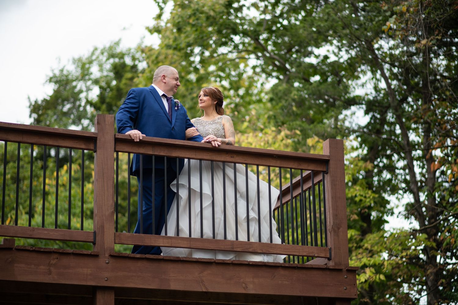 Newlyweds share an intimate moment on a wooden deck overlooking trees.