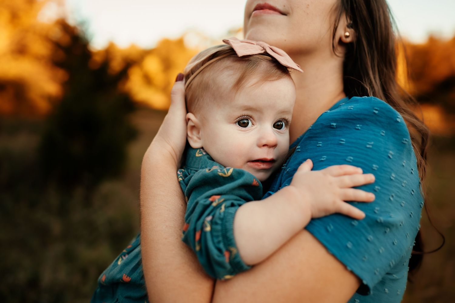 Parent holds baby in blue shirt against autumn backdrop during sunset photoshoot.