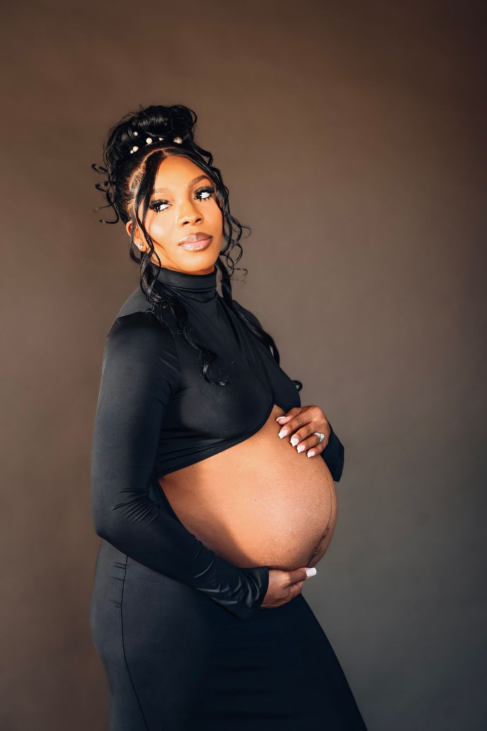Pregnant Black woman in black turtleneck crop top and skirt cradling baby bump, studio portrait on brown background.