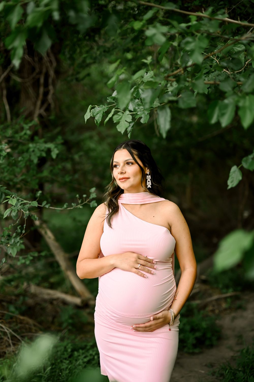 Pregnant woman in pink one-shoulder maternity dress posing outdoors among green trees.