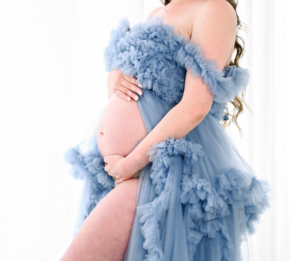 Pregnant woman in elegant off-shoulder dusty blue ruffled tulle maternity gown posing for a photoshoot.