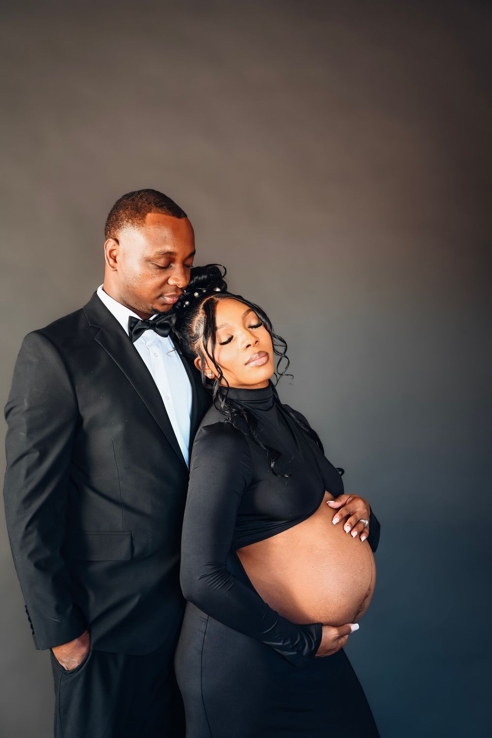 Elegant couple in black formal attire posing for maternity photo, man in tuxedo embracing pregnant woman against gray backdrop.