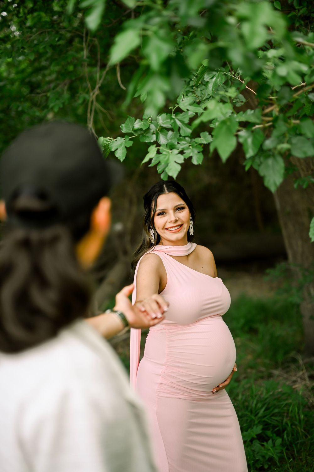 Smiling pregnant woman in pink maternity gown holds hands with partner outdoors under green trees.