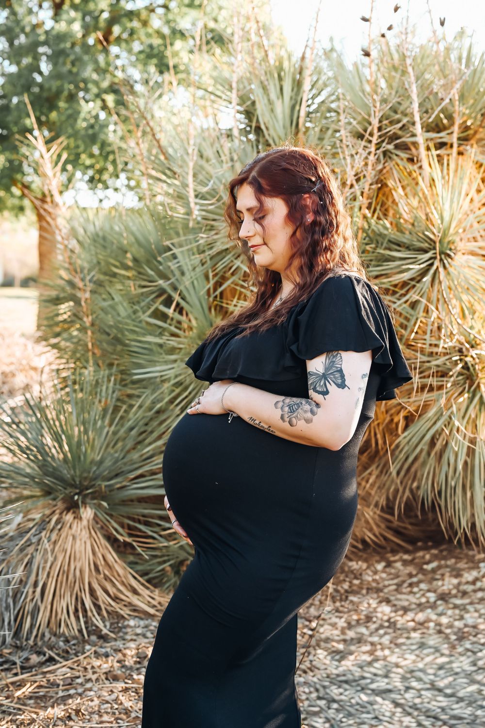 Pregnant woman in black dress holding baby bump outdoors near yucca plants, showing butterfly tattoo on arm.