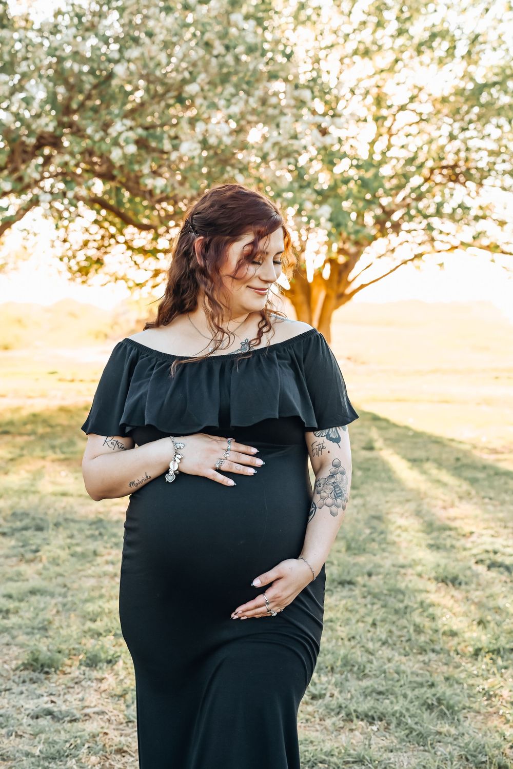 Pregnant woman in black off-shoulder gown cradling baby bump outdoors at golden hour near blooming tree.