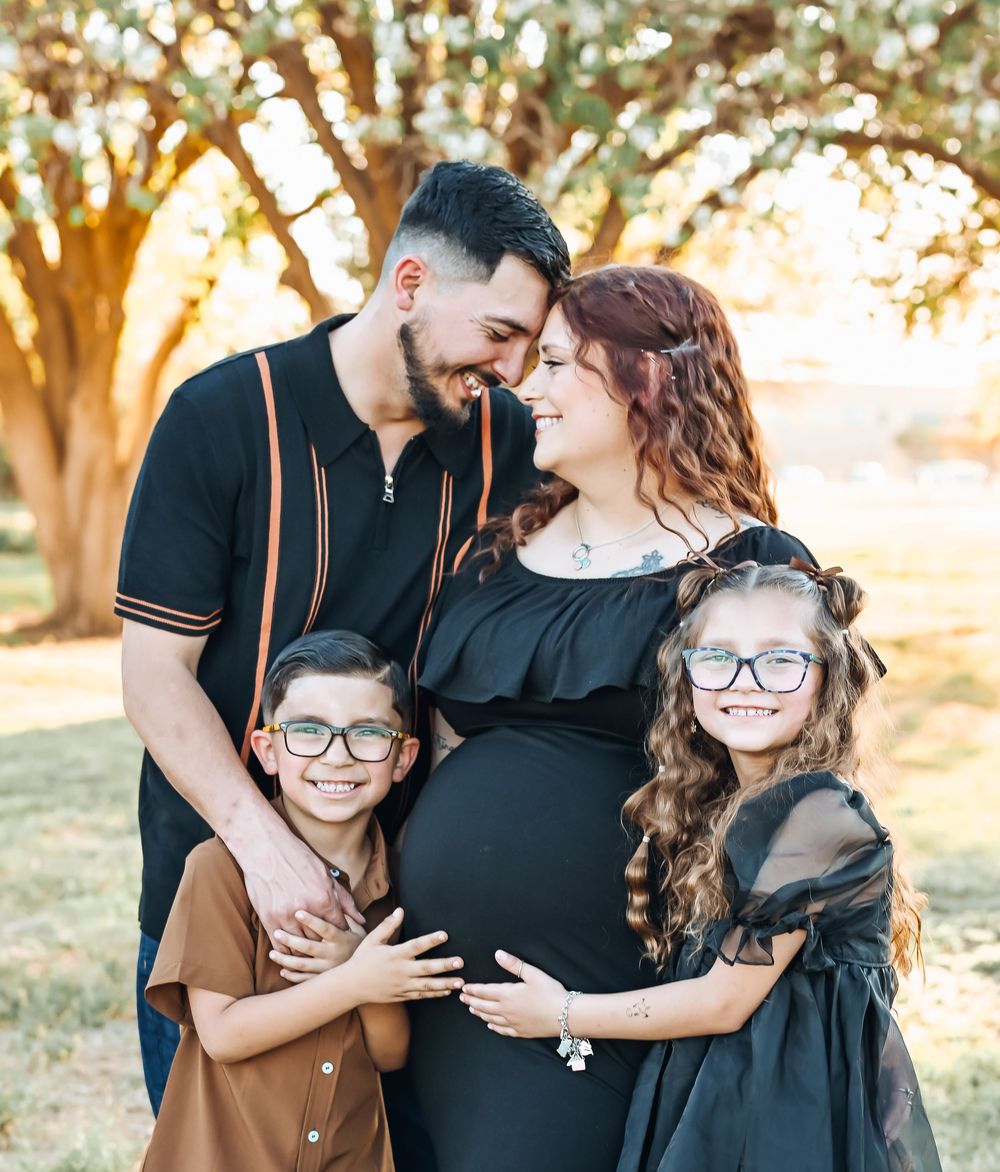 Pregnant woman poses with husband and two children wearing glasses outdoors during golden hour maternity photo session.