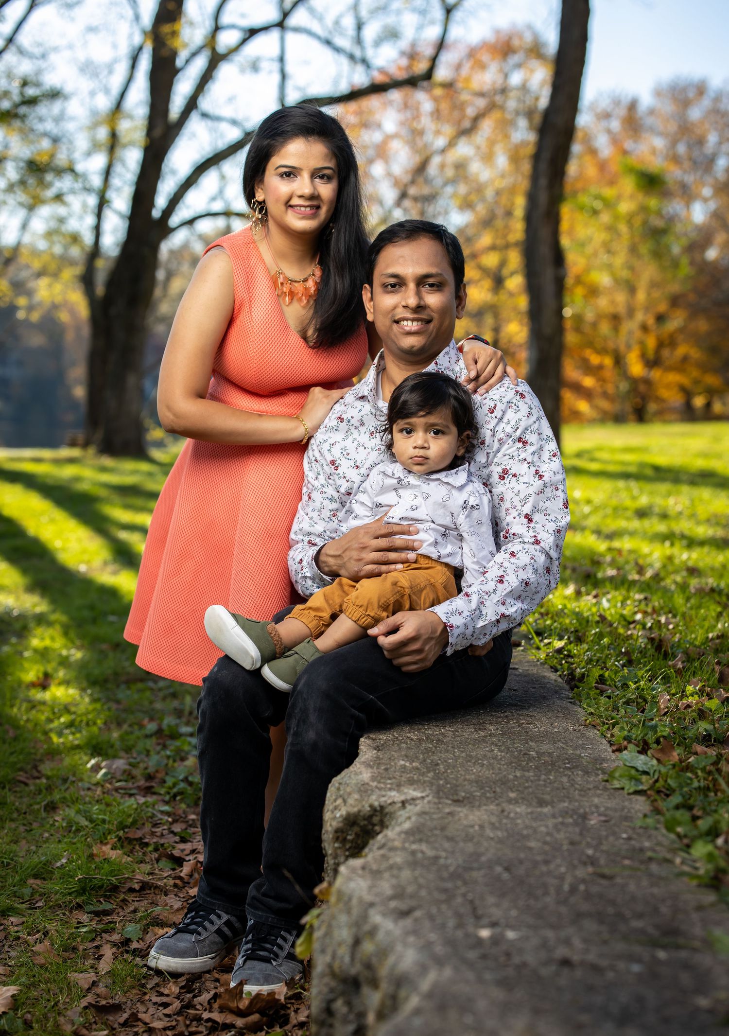 Family of three poses on stone ledge during autumn outdoor photo session.