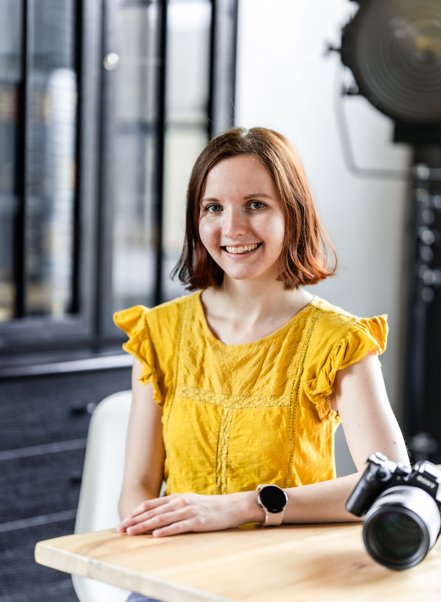 A person in a yellow ruffled blouse sitting at a table with camera equipment.