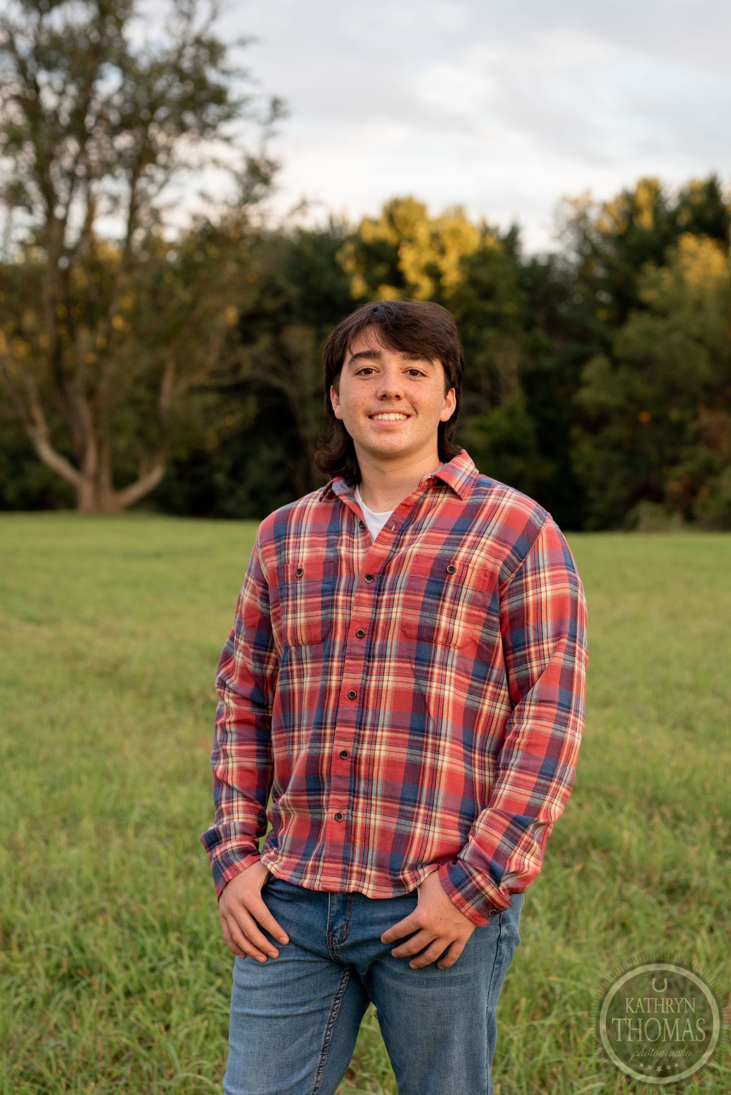 Person in red plaid flannel shirt smiles while standing in sunny meadow.