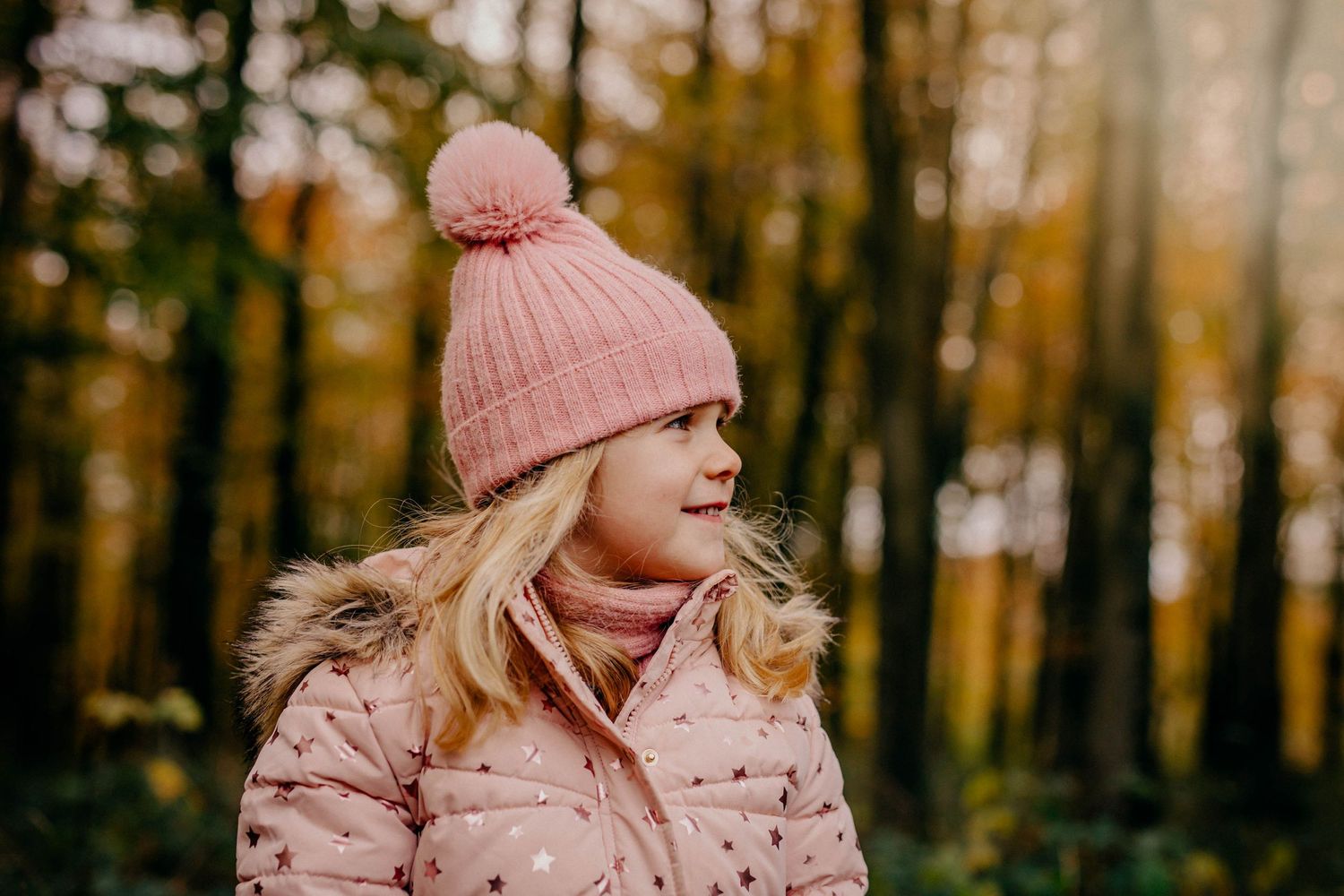 Young girl in a pink bobble hat and coat smiling in the autumn woods