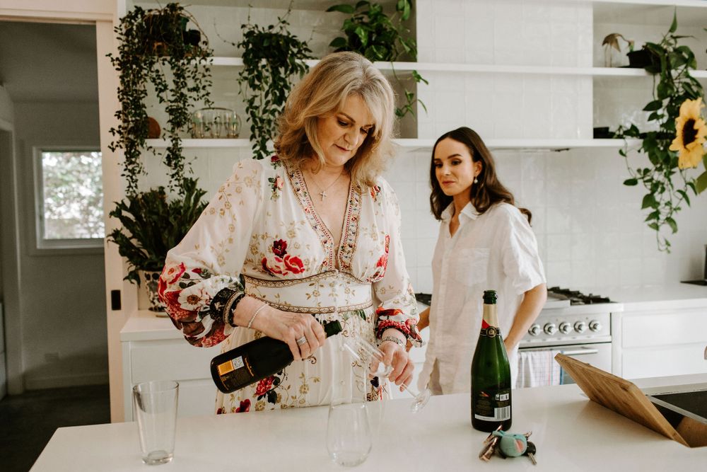 Women in floral dresses preparing beverages in bright modern kitchen with hanging plants.