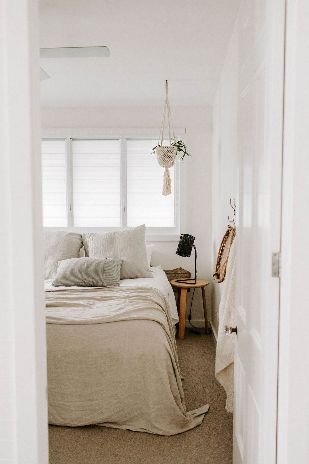 Minimalist bedroom interior with white linens, wooden furniture, and a hanging plant in a macrame holder.