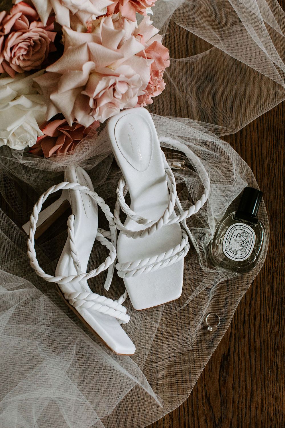 Wedding accessories including strappy white sandals and pink roses with tulle veil on wooden surface.