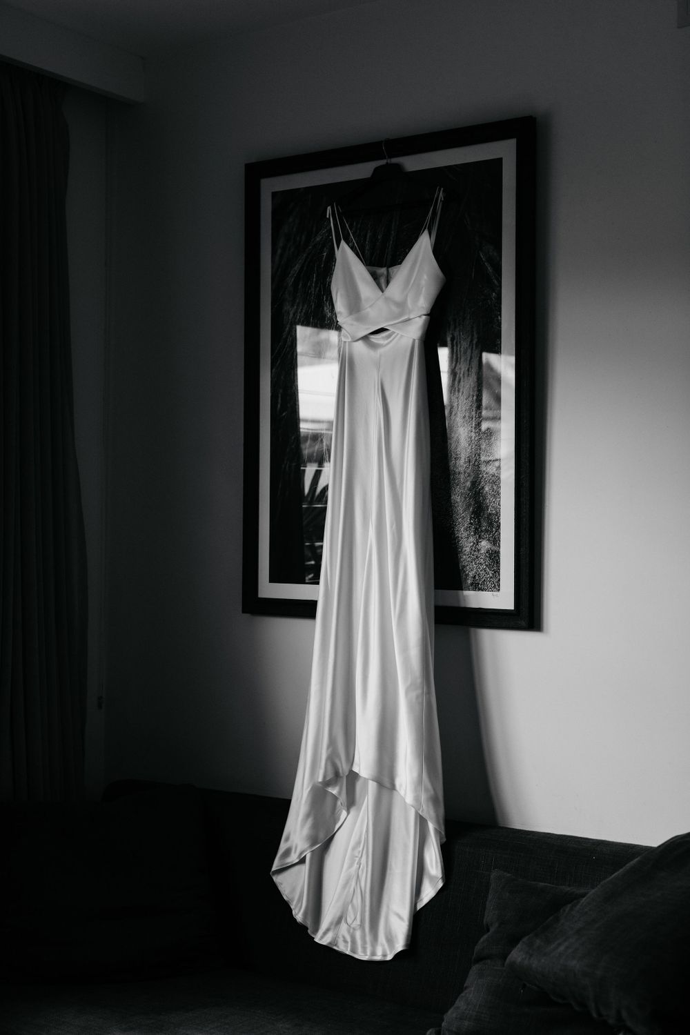 A white silk wedding dress hanging against a dark wall with framed artwork.