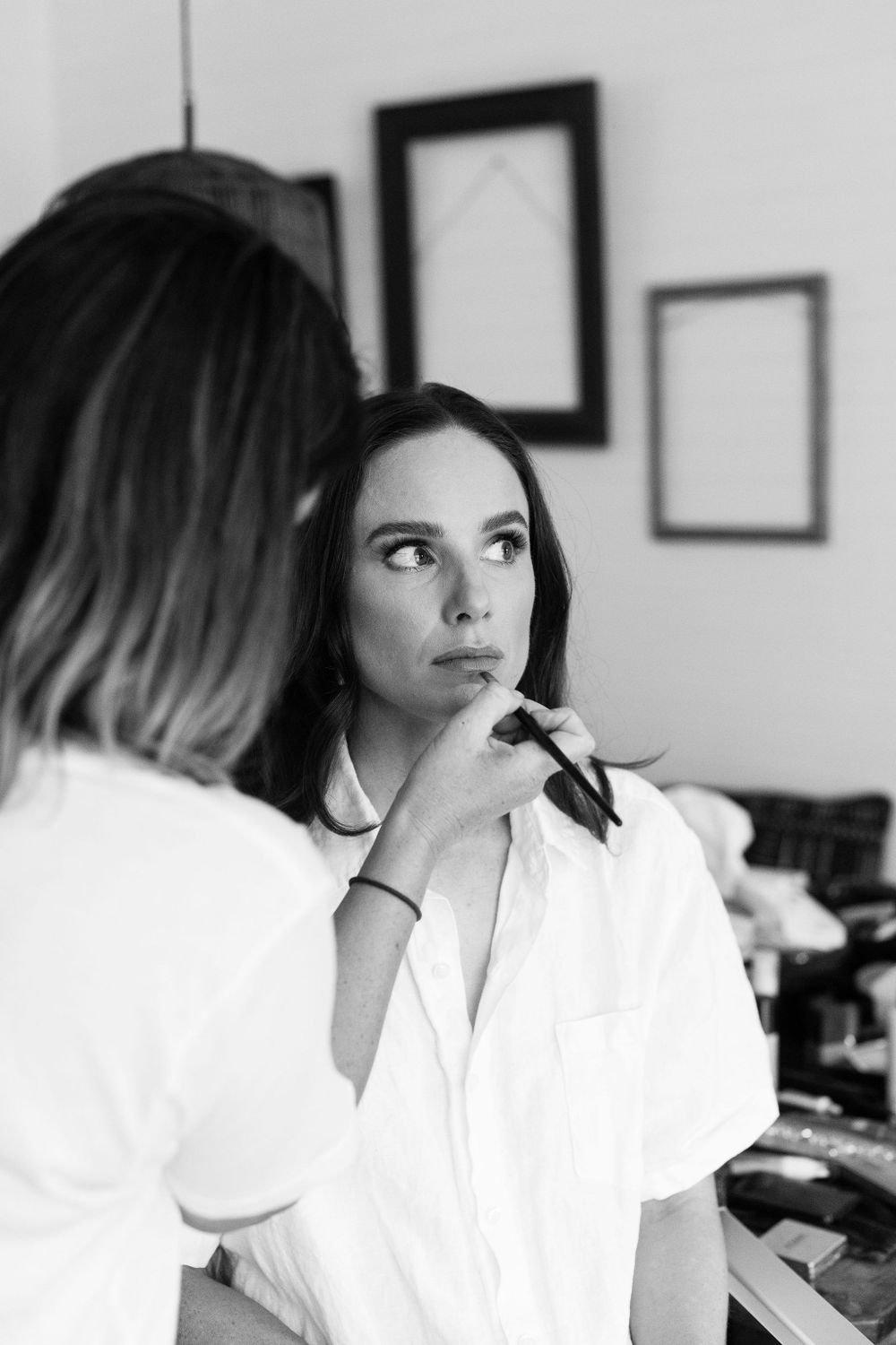 Woman getting makeup applied while sitting in white dress against neutral wall with frames.