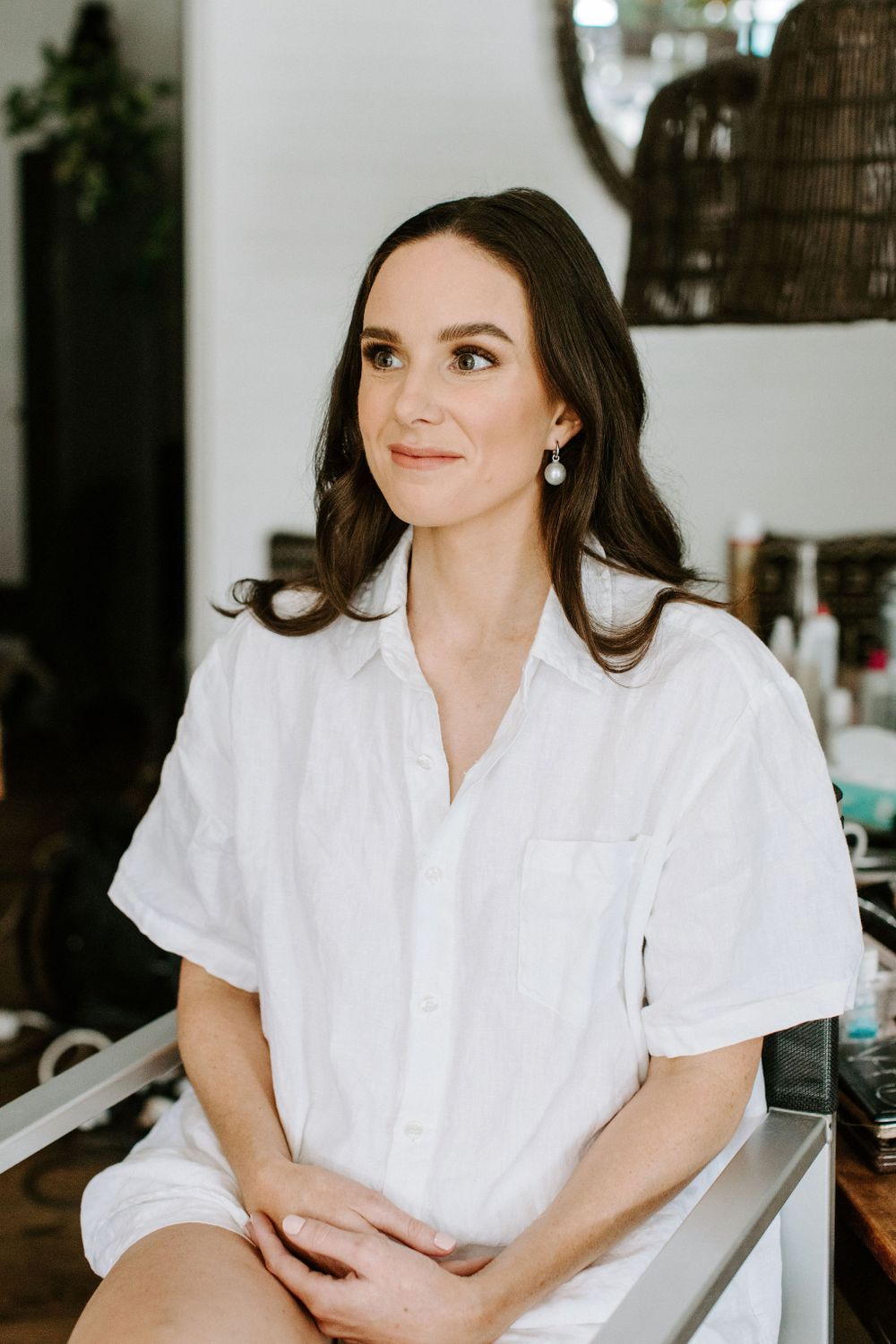 Woman in crisp white button-down shirt seated elegantly during beauty preparation.
