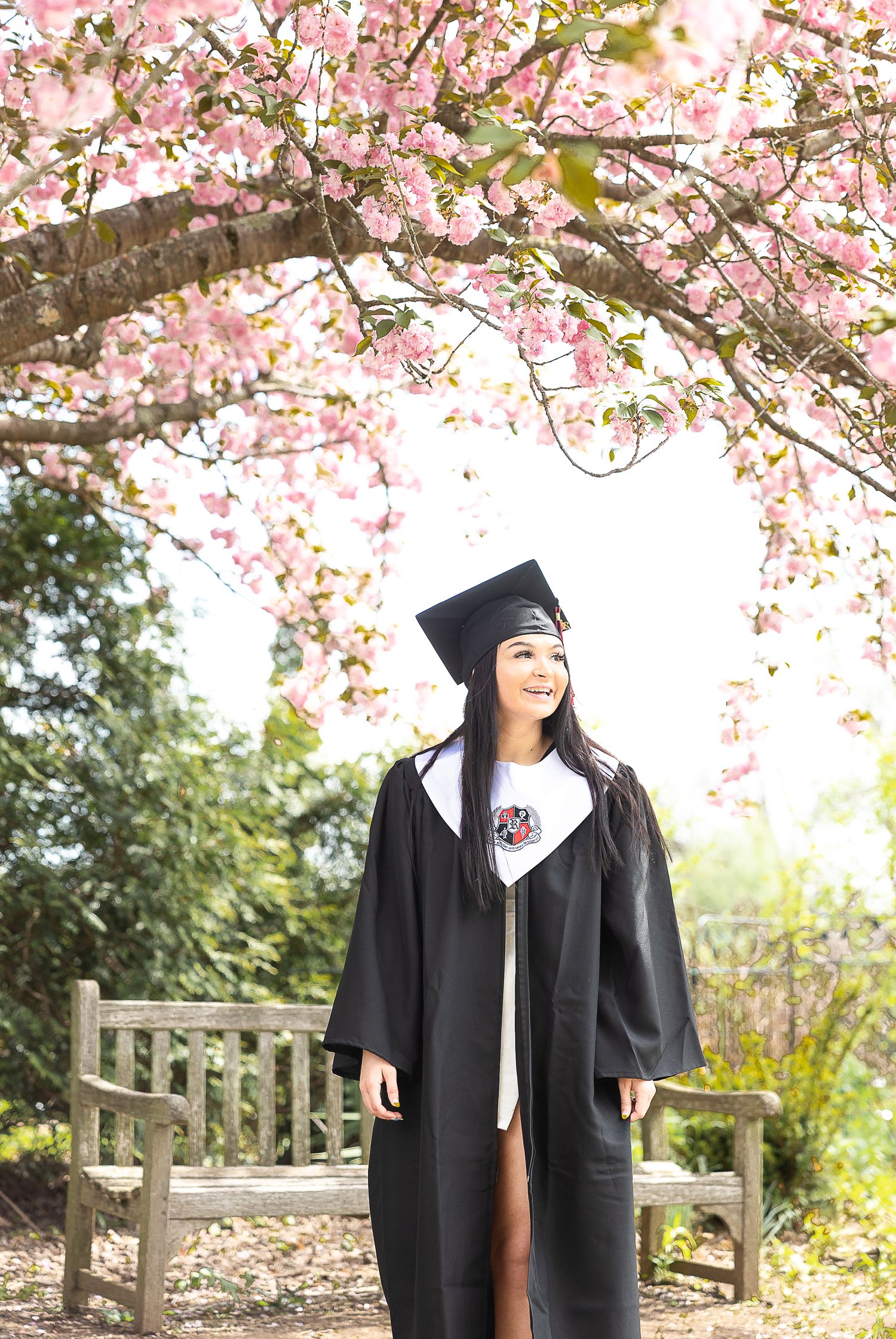 Graduate in traditional black gown stands under flowering cherry blossom trees.