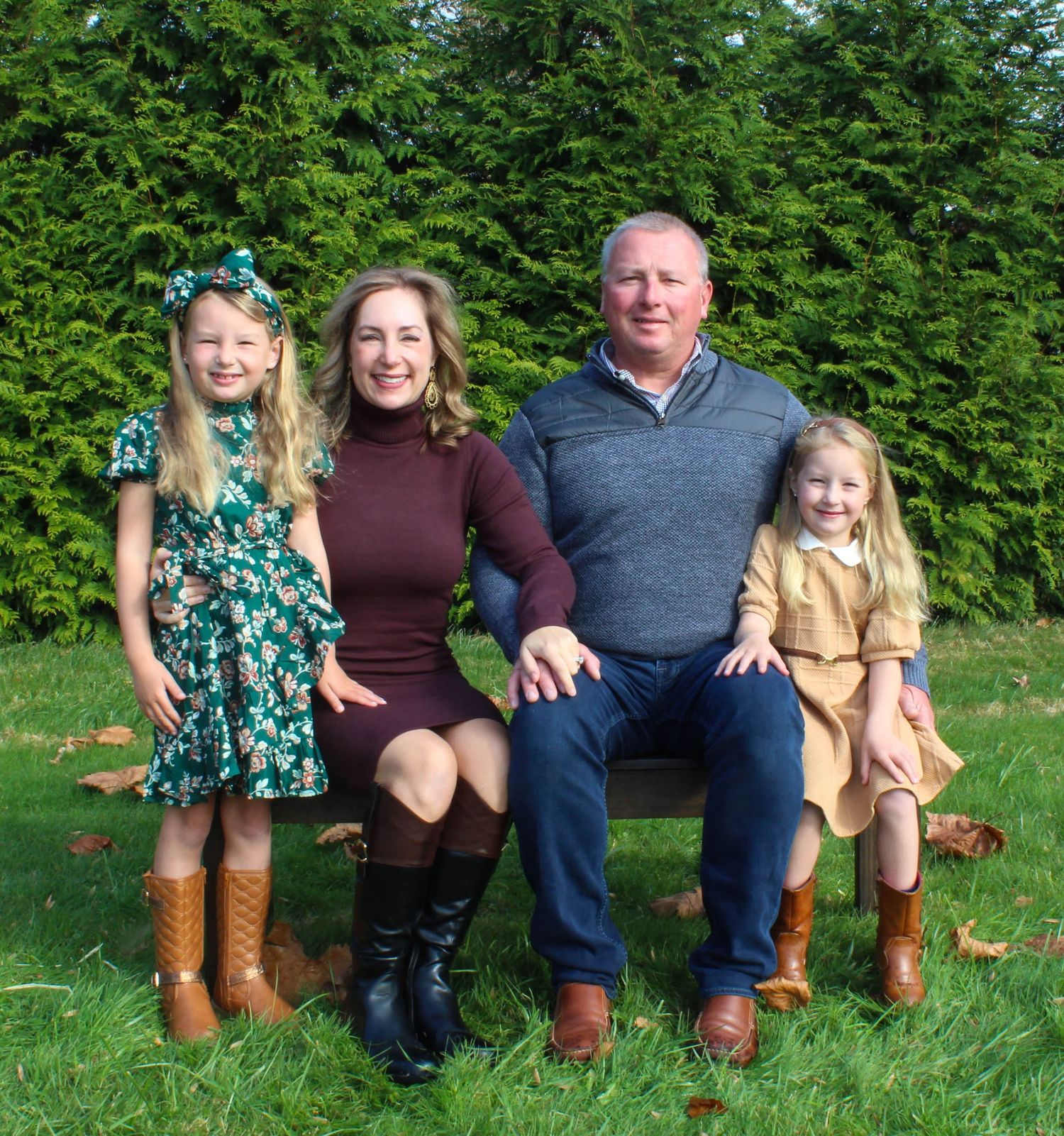 Family group posed on grass with evergreen hedge backdrop wearing coordinated fall outfits.