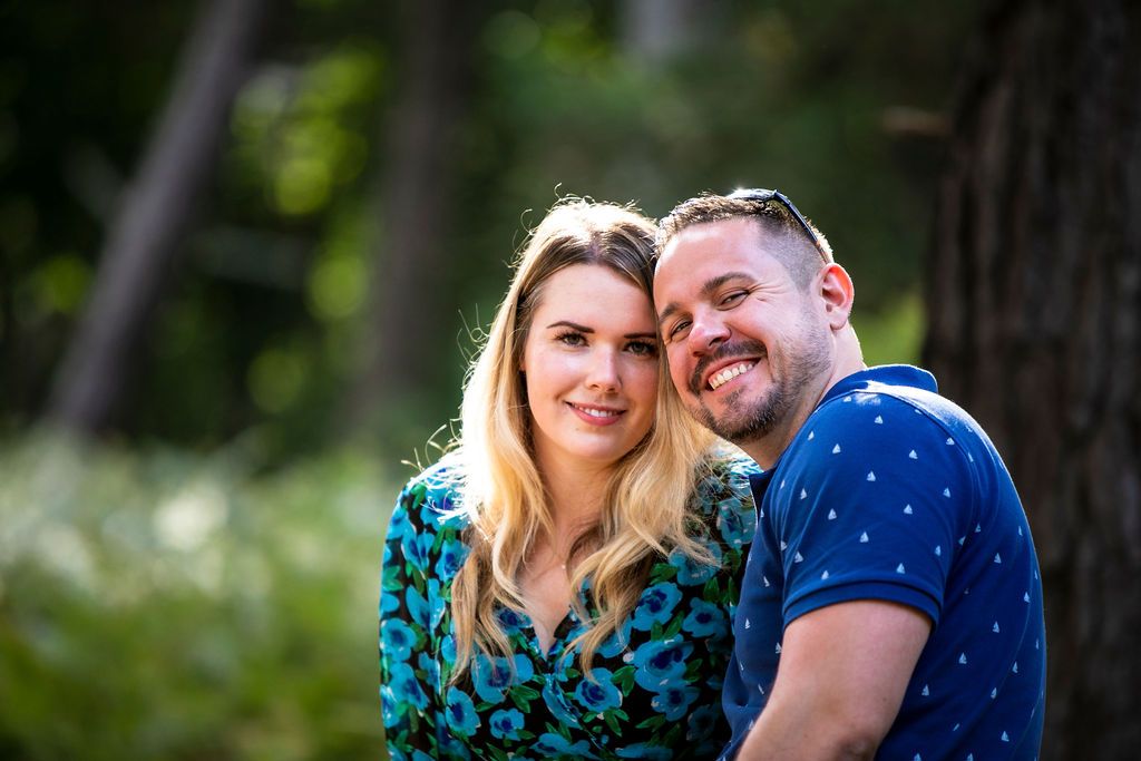 A close-up outdoor portrait of a couple in casual attire smiling against a natural background.