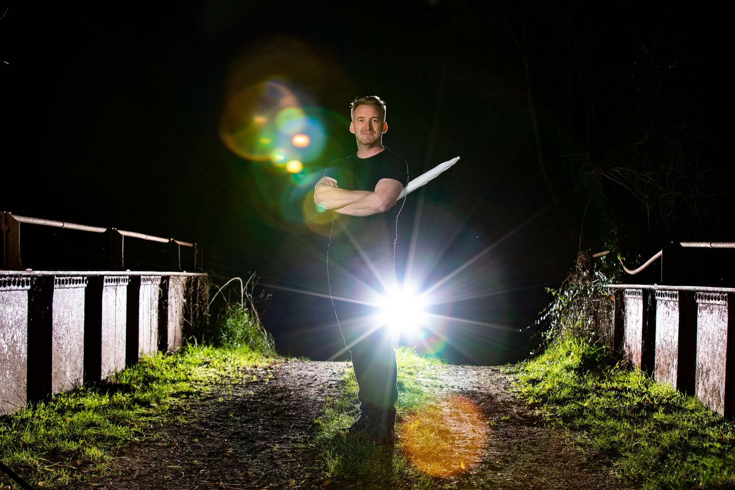 Photographer with flashlight standing on dark path between railings at night.