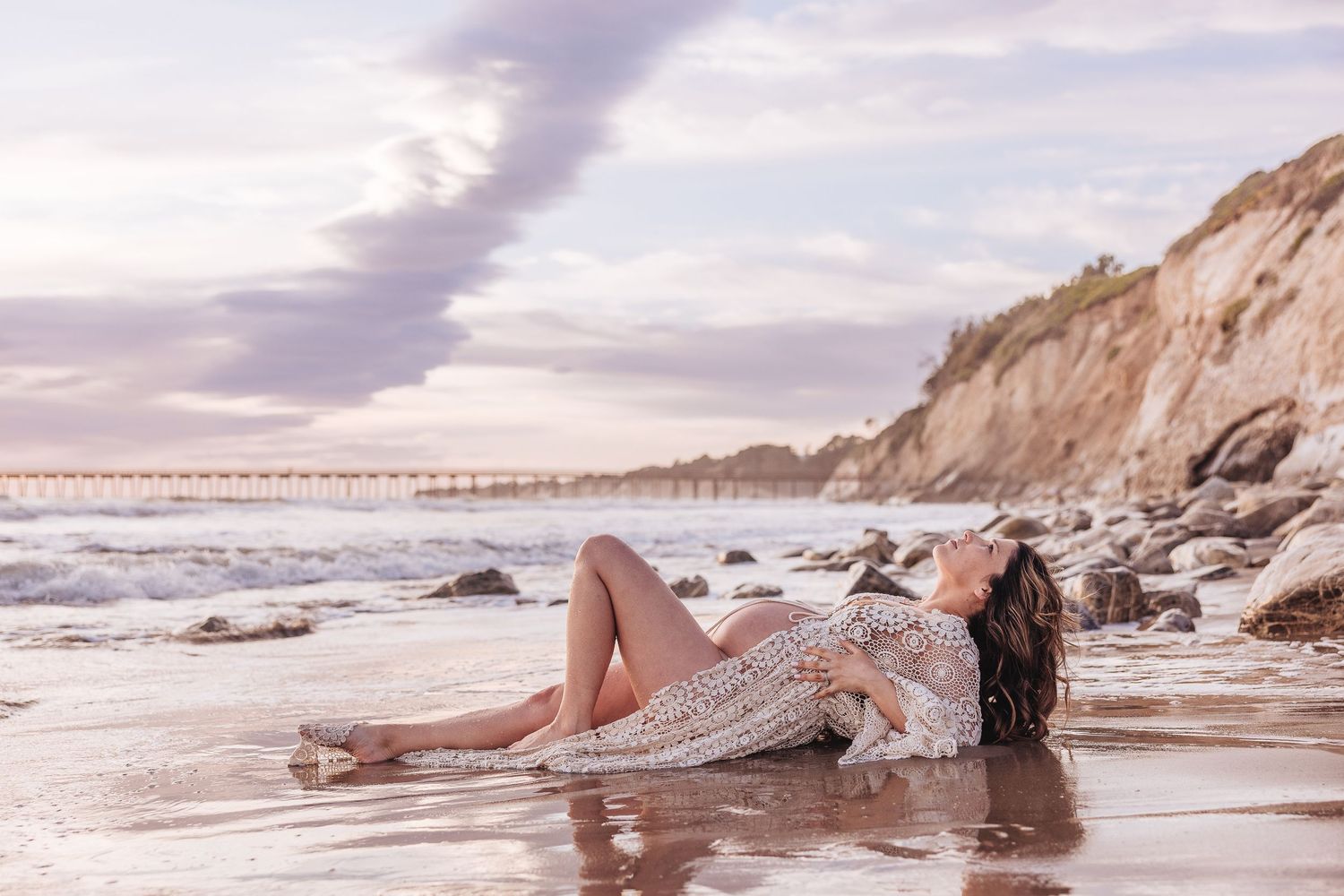 A dreamy beachside maternity photo with waves and cliffs at sunset.