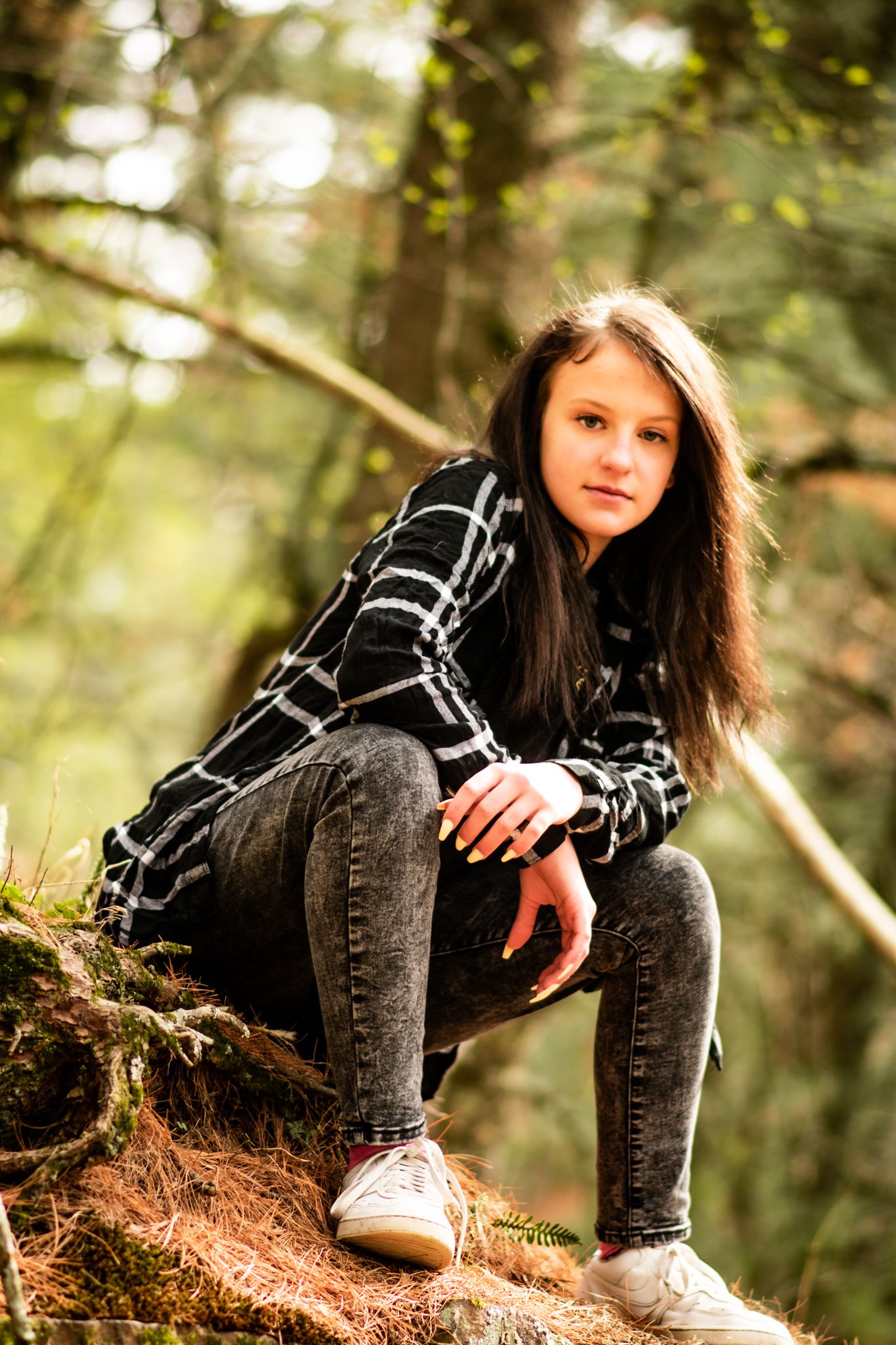 A person in a plaid shirt and jeans sits casually on a mossy slope in a sunlit forest.
