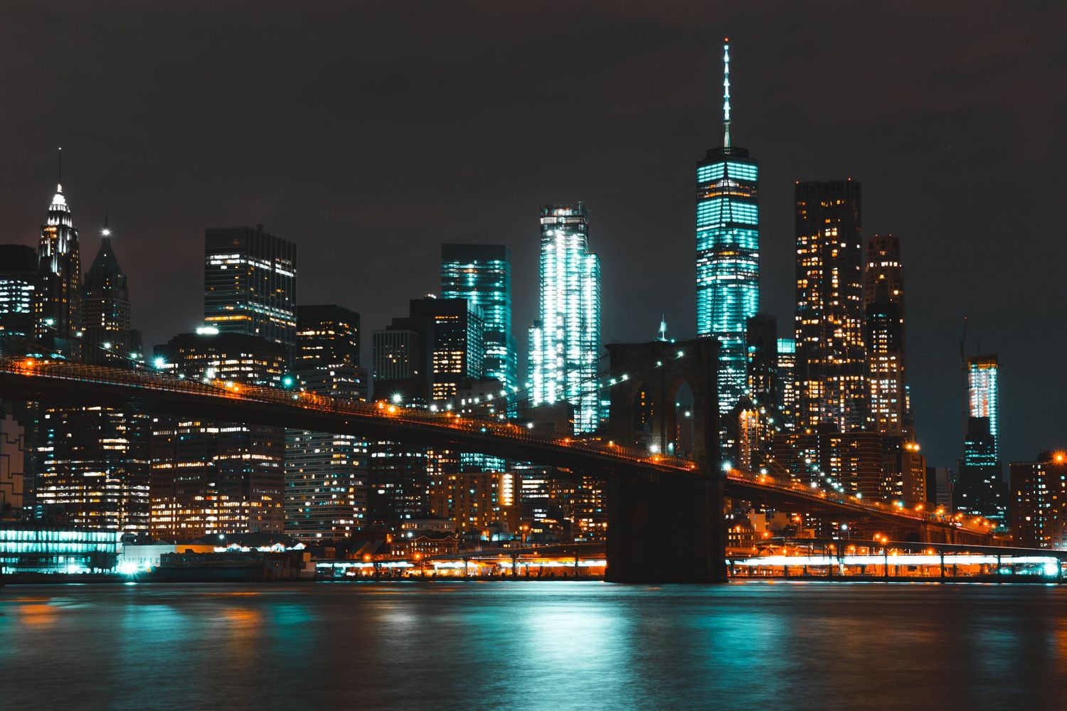 Brooklyn Bridge and Lower Manhattan skyline illuminated at night reflecting on the East River.