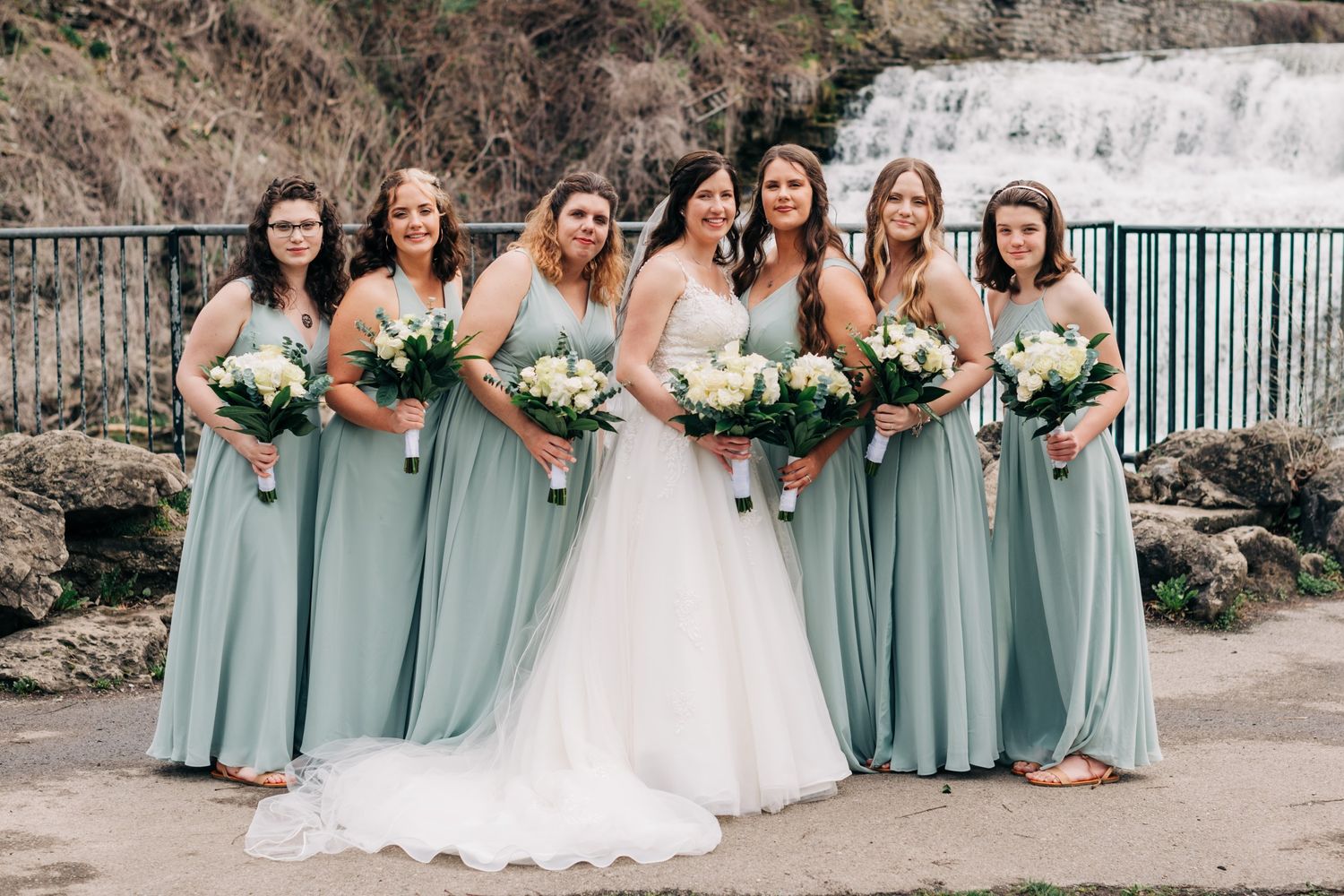 bride and bridesmaids in eucalyptus green dresses pose in front of a waterfall