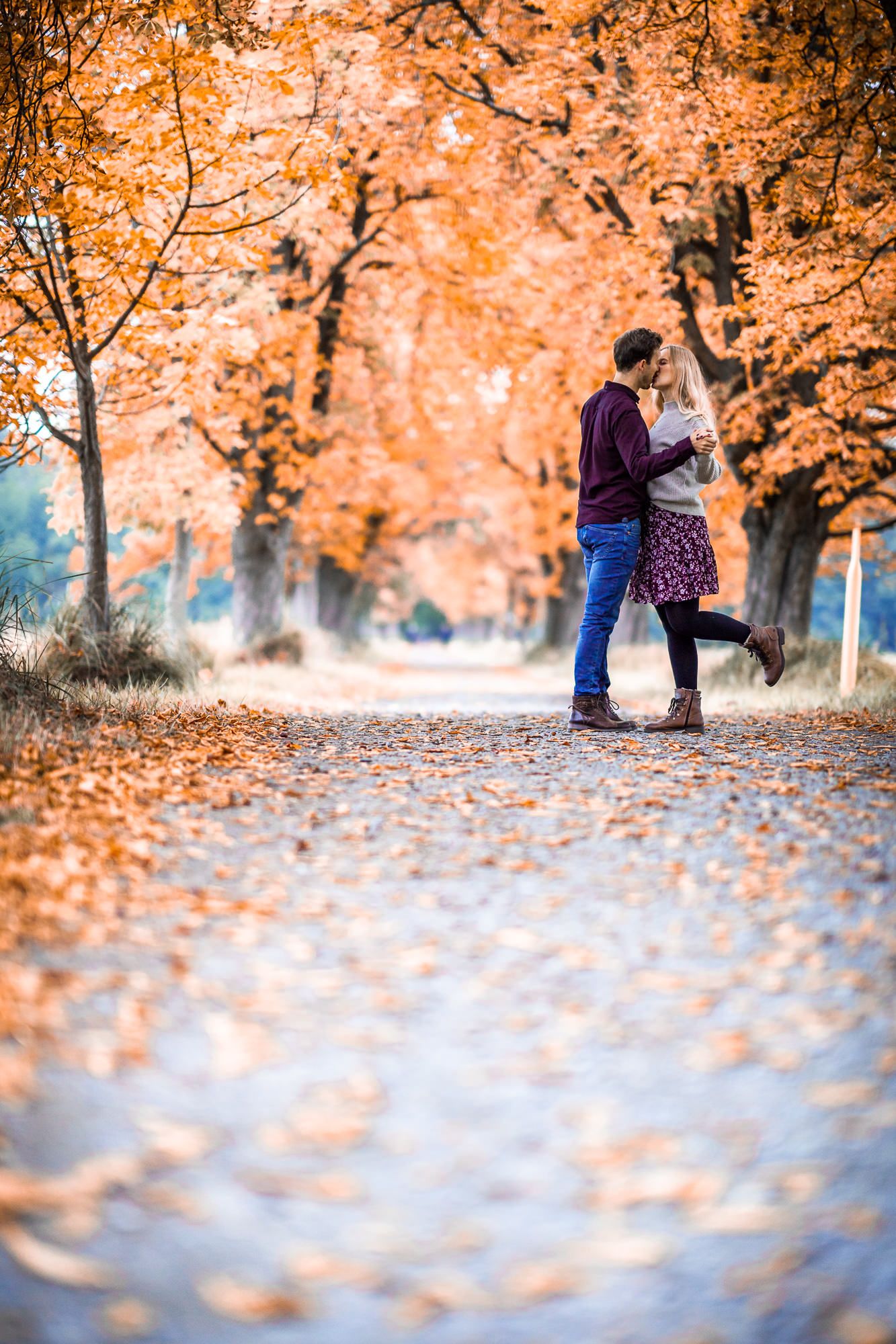 A couple share an intimate moment on an autumn path covered in fallen orange leaves.