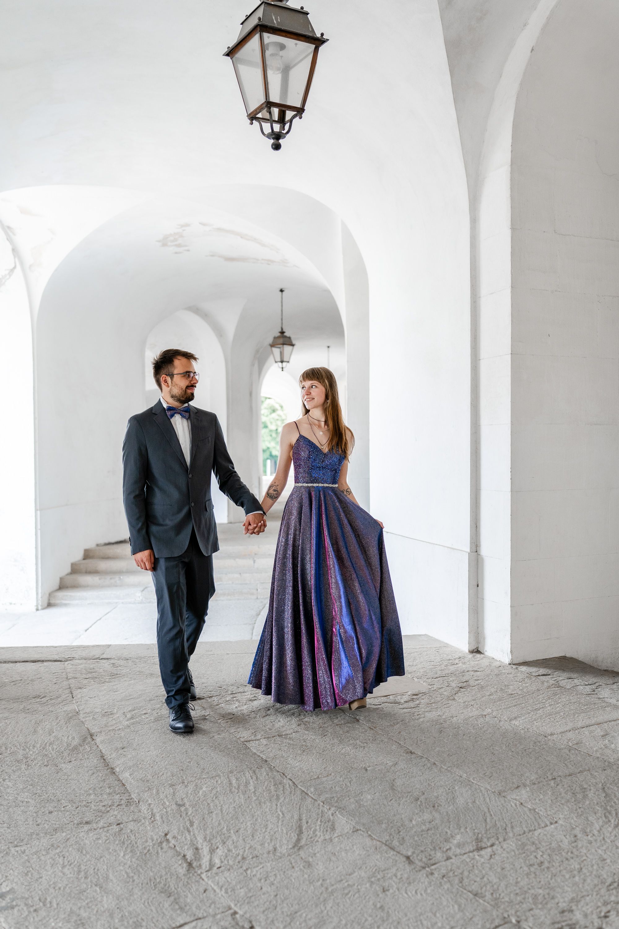 Elegant couple in formal wear walking through white arched hallway.