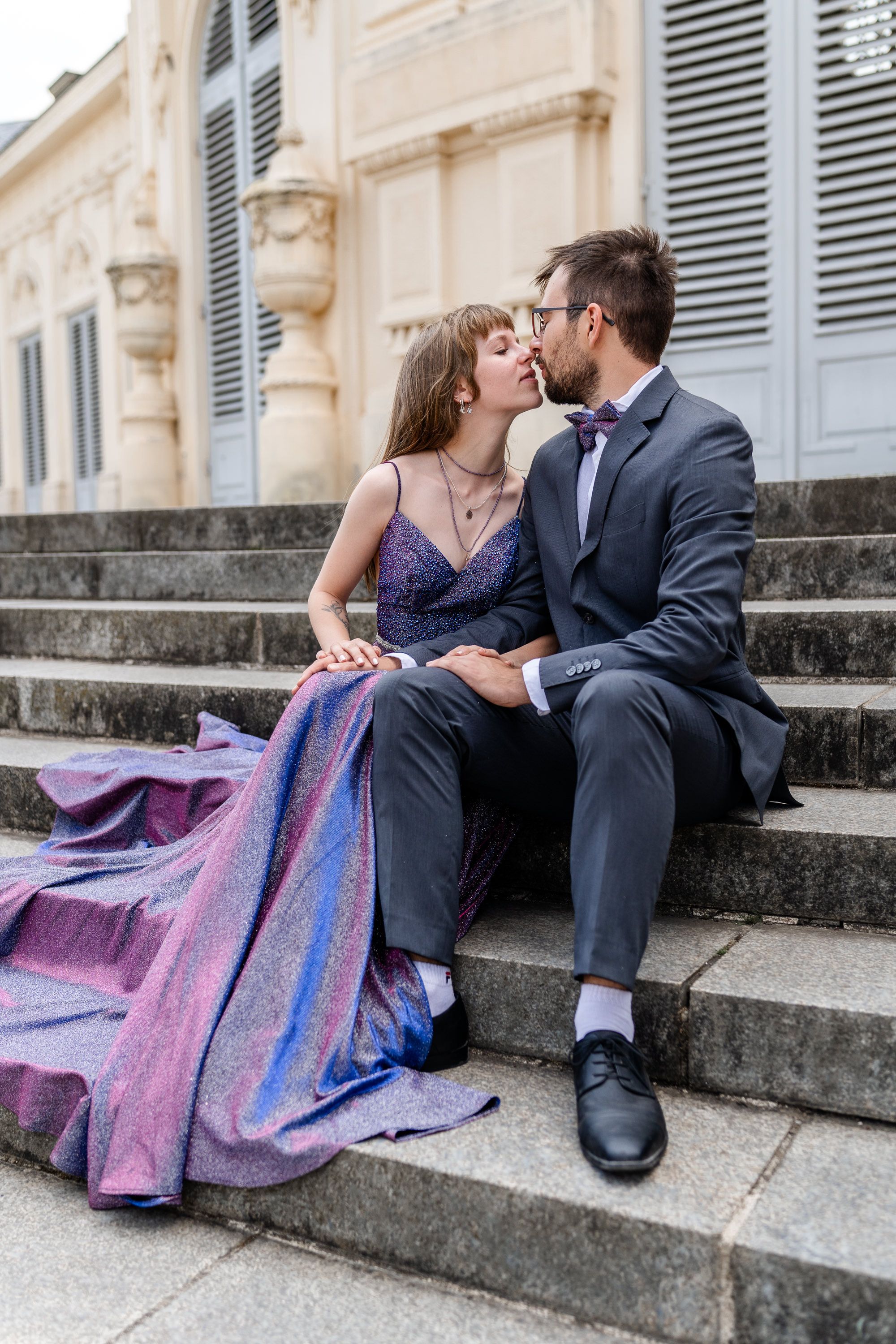 Romantic couple sitting on stone steps, wearing formal attire with a sparkly purple evening gown and dark suit.