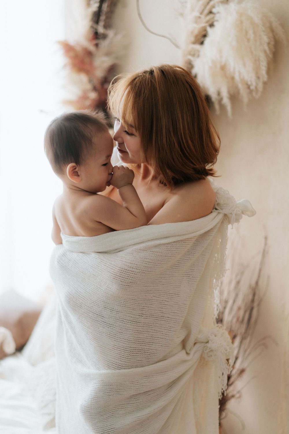 A gentle moment captured between parent and infant wrapped in soft white fabric by a window.