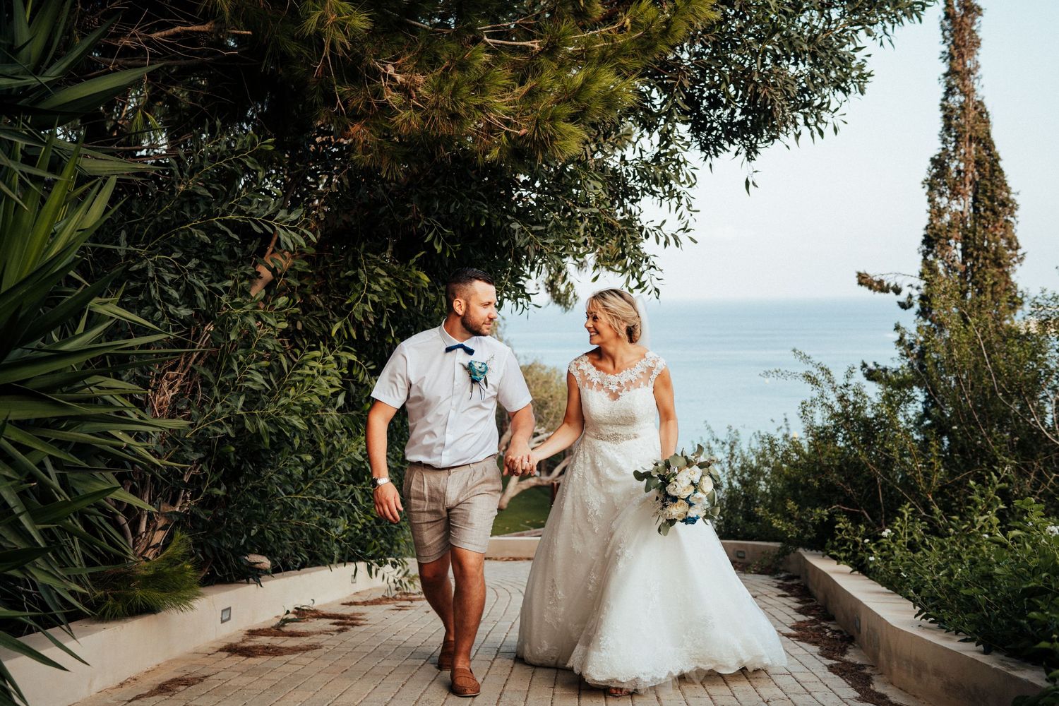 The wedding couple walk hand in hand along a scenic coastal path surrounded by Mediterranean vegetation.