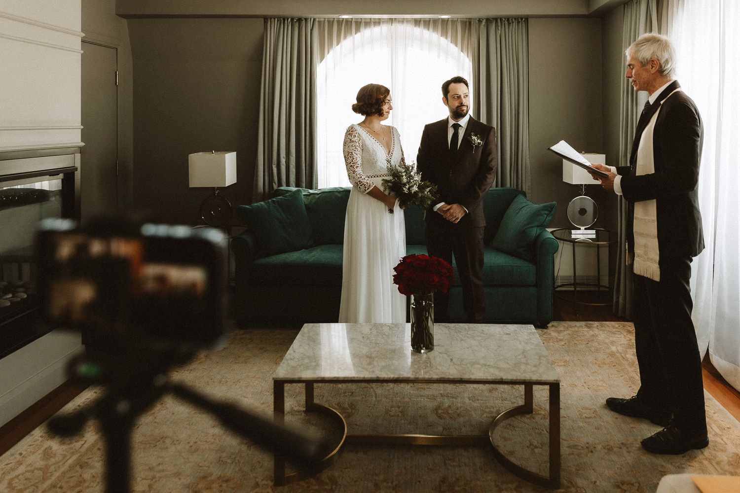 An intimate indoor wedding ceremony takes place in a cozy room with a camera in the foreground.