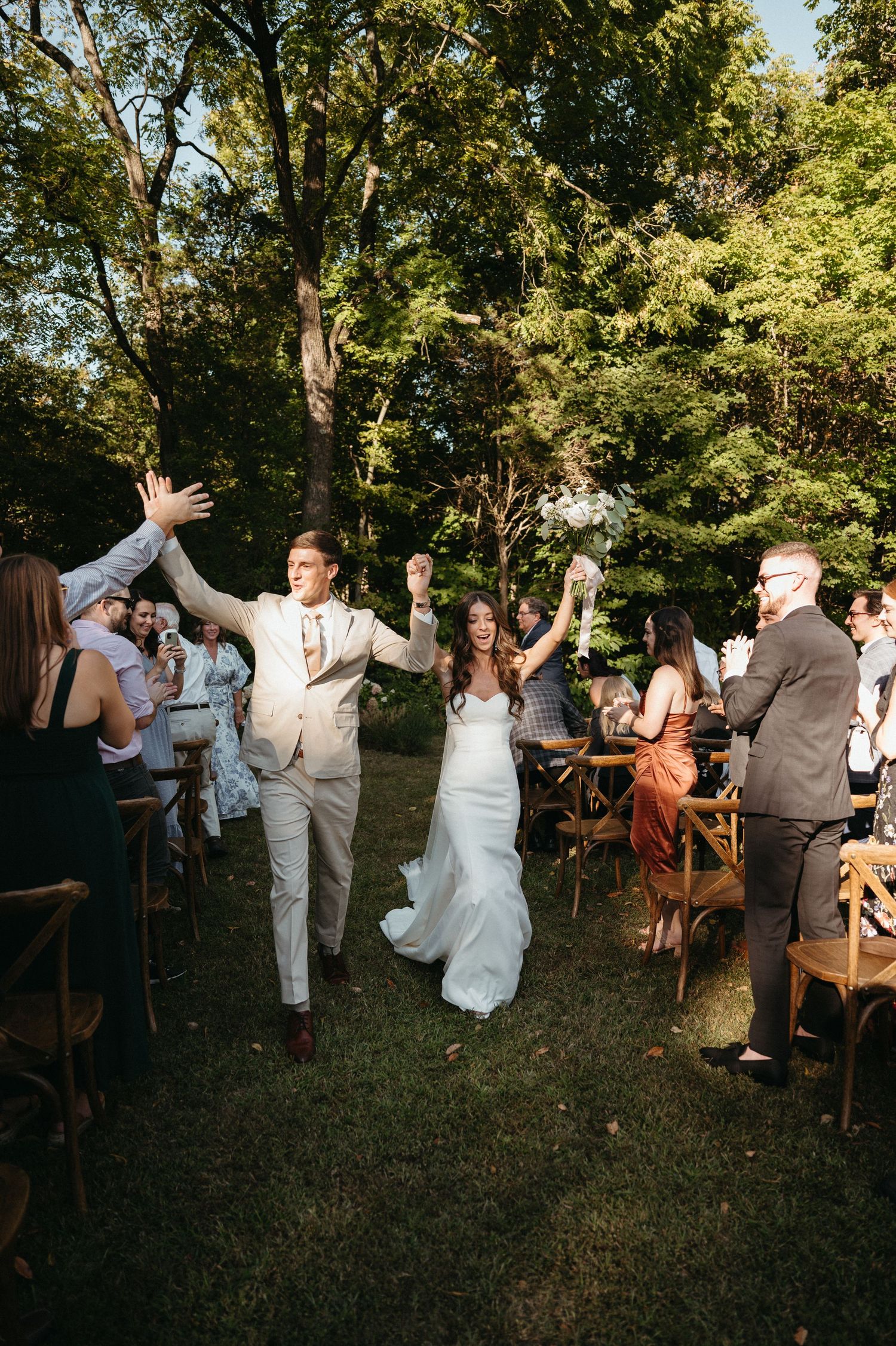 Wedding guests cheer as newlyweds walk down the aisle in an outdoor garden ceremony.